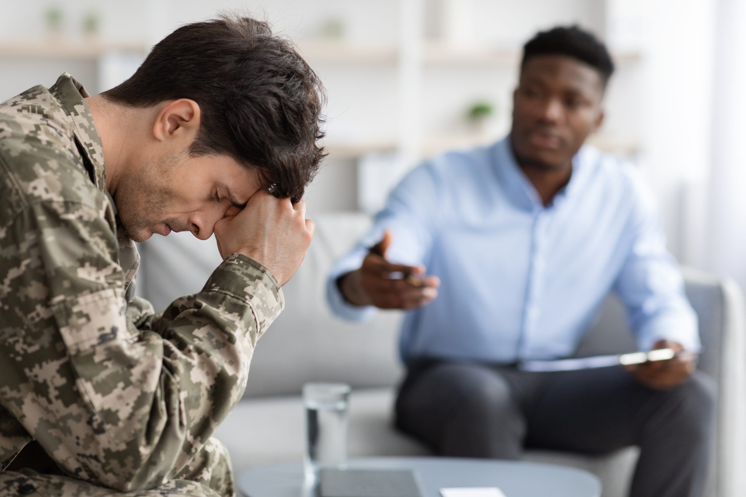 Individual Counseling: Distressed young man in uniform soldier attending therapy session with african american psychiatrist, sitting on chair and touching his head, psychological trauma in military personnel concept